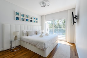 Een moderne slaapkamer met een groot bed, een nachtkastje, een televisie aan de muur en een raam met gordijnen.