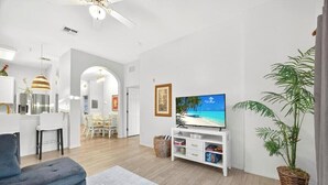 Una sala moderna con una televisión de pantalla plana, un sofá gris, un comedor con sillas blancas y una planta grande en maceta.