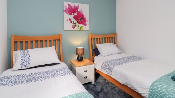 A twin room with two beds, a nightstand, a lamp, and a floral artwork on the wall. - Mayfly Lodge (Bodmin)