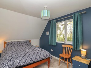 A bedroom with a bed, a chair, a nightstand, a lamp, and a window with curtains. - Mayfly Lodge (Bodmin)