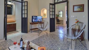 Ein offener Wohnbereich mit Fernseher, zwei weiße Stuhlgestelle, einem Esstisch mit Flasche und Gläsern sowie einem gemusterten Boden.