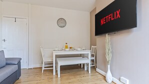 Ein Wohnzimmer mit einem weißen Sofa, einem Esstisch gedeckt mit Tellern und Gläsern, einer Uhr an der Wand und einem Fernseher, der 'NETFLIX' zeigt.