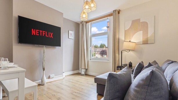 Ein Wohnzimmer mit einem Flachbildfernseher, der 'NETFLIX' anzeigt, einem grauen Sofa, einem weißen Beistelltisch und einem Fenster mit Vorhängen.