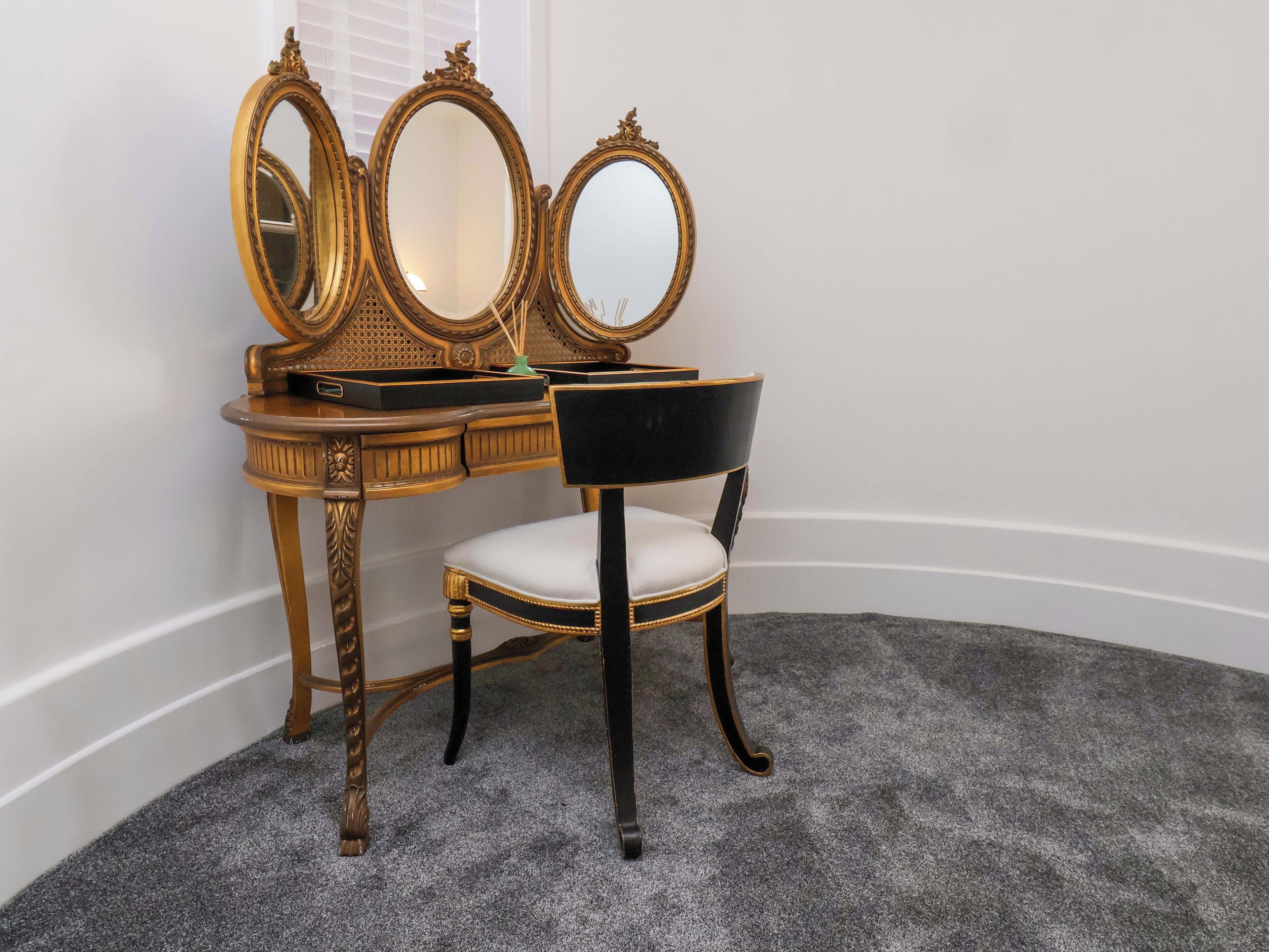 A corner room with a vanity table, three mirrors, a chair, and a carpeted floor.