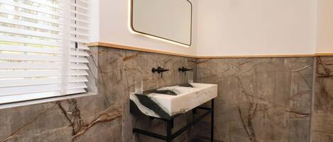 Un baño con pared de mármol, un lavamanos y una ventana con persianas.