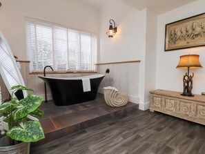 Ein Badezimmer mit einer freistehenden Badewanne, einem Fenster mit Jalousien, einer wandmontierten Leuchte, einem dekorativen Teppich und einem Schrank mit einer Lampe.