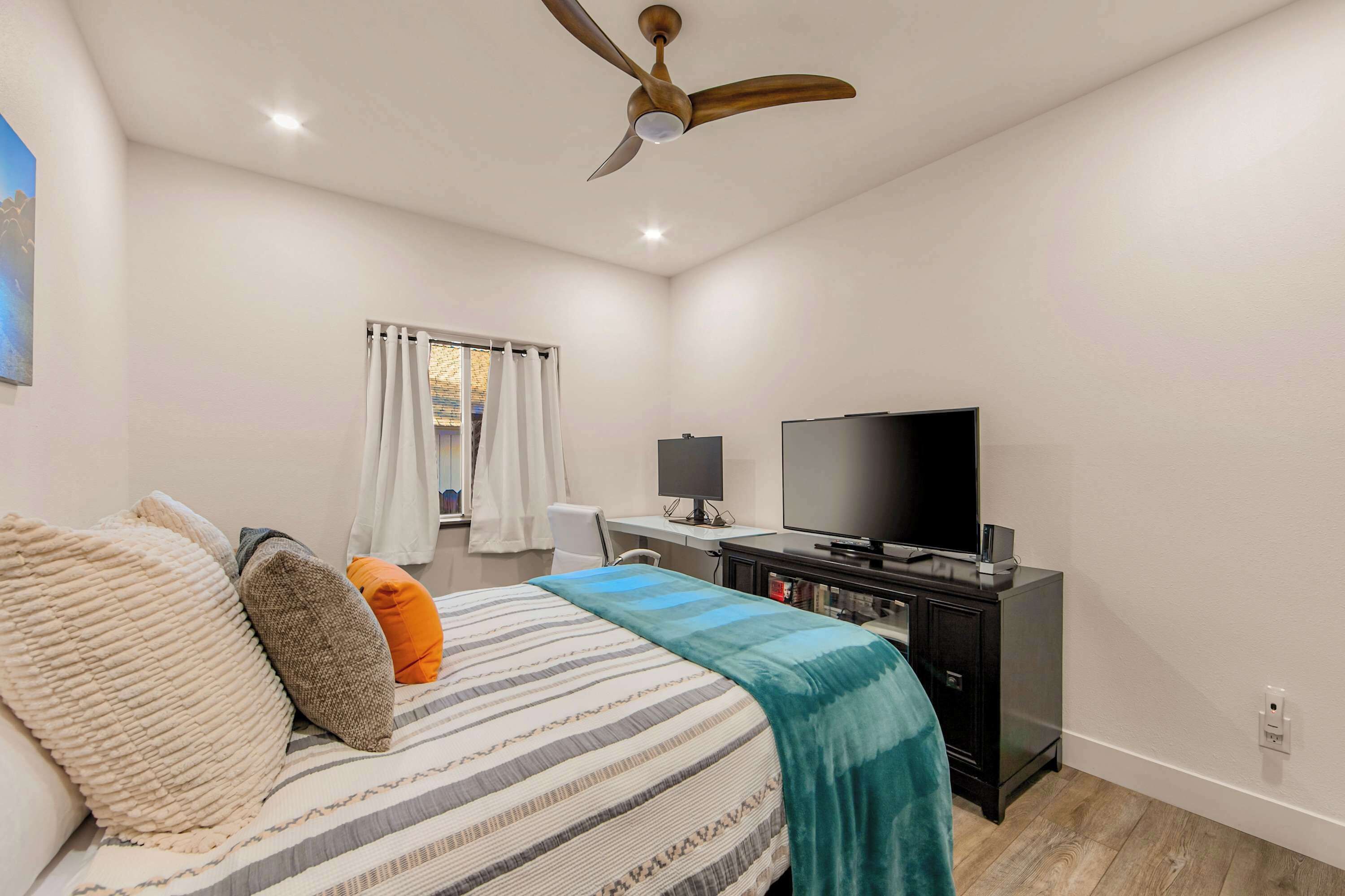 Un dormitorio con cama, almohadas, televisor, escritorio y ventilador de techo.