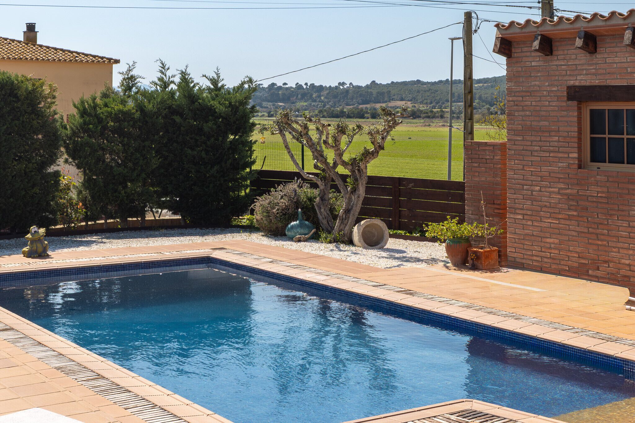 A swimming pool with a tiled edge, surrounded by a brick building and a fenced area with trees and plants.