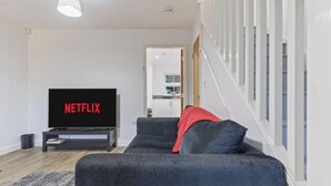 A modern living room with a flat-screen TV, a dark sofa, and a staircase leading to an upper floor. - Amy`s House - close to the Peak District! (Alton Towers & Staffordshire Villages)