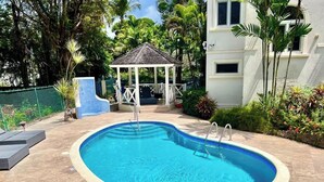Una piscina con un pabellón, rodeada de exuberante follaje y un edificio blanco.