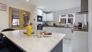A modern kitchen with white cabinets, a dining table with a yellow bottle, and a bowl of fruit. - Syston Townhouse With parking (Leicester)