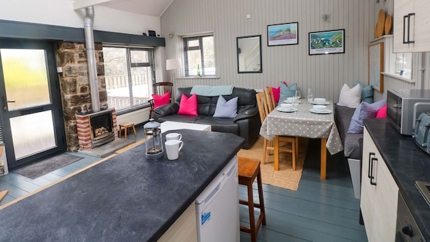 A compact kitchen with a central island, a dining area with a table set for two, and a living area with a sofa and chairs. - Brewery Cottage (Saundersfoot)