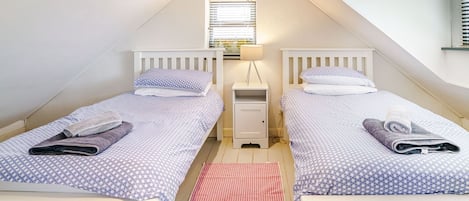 A small, well-lit attic bedroom with two single beds, a nightstand, a lamp, and a red mat on the floor.