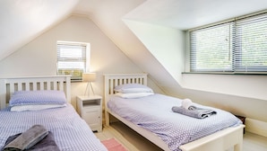 A bedroom with two beds, a window with blinds, a bedside table with a lamp, and a red rug. - Brewery Cottage (Saundersfoot)