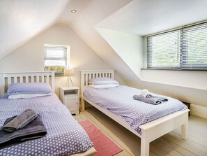 A bedroom with two beds, a window with blinds, a bedside table with a lamp, and a red rug. - Brewery Cottage (Saundersfoot)
