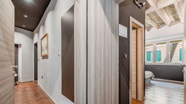 Un corridoio con pavimento in legno, una porta che dà su una stanza con un letto e una finestra con persiane verdi.