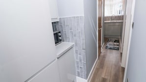 Un couloir étroit avec un coin cuisine comprenant des meubles blancs, un pare-feu en carrelage et un plancher en bois.