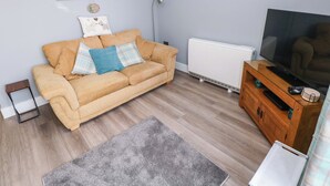 Un salon avec un canapé beige, un tapis gris, un meuble TV en bois et un radiateur blanc.