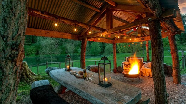 A rustic wooden pavilion with a fire pit, lanterns, and a bench.