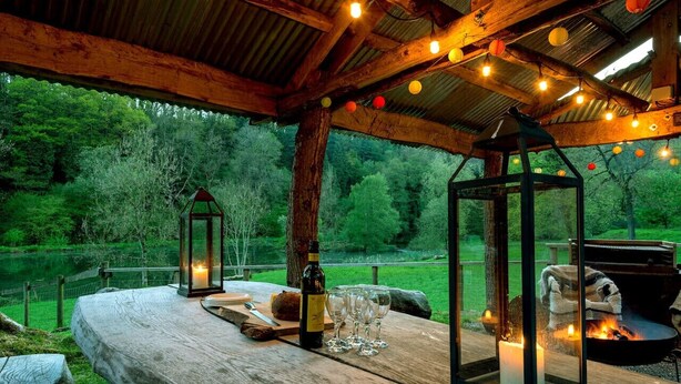 A rustic wooden table with a bottle, glasses, and a candle, set under a wooden roof with string lights, overlooking a green landscape.