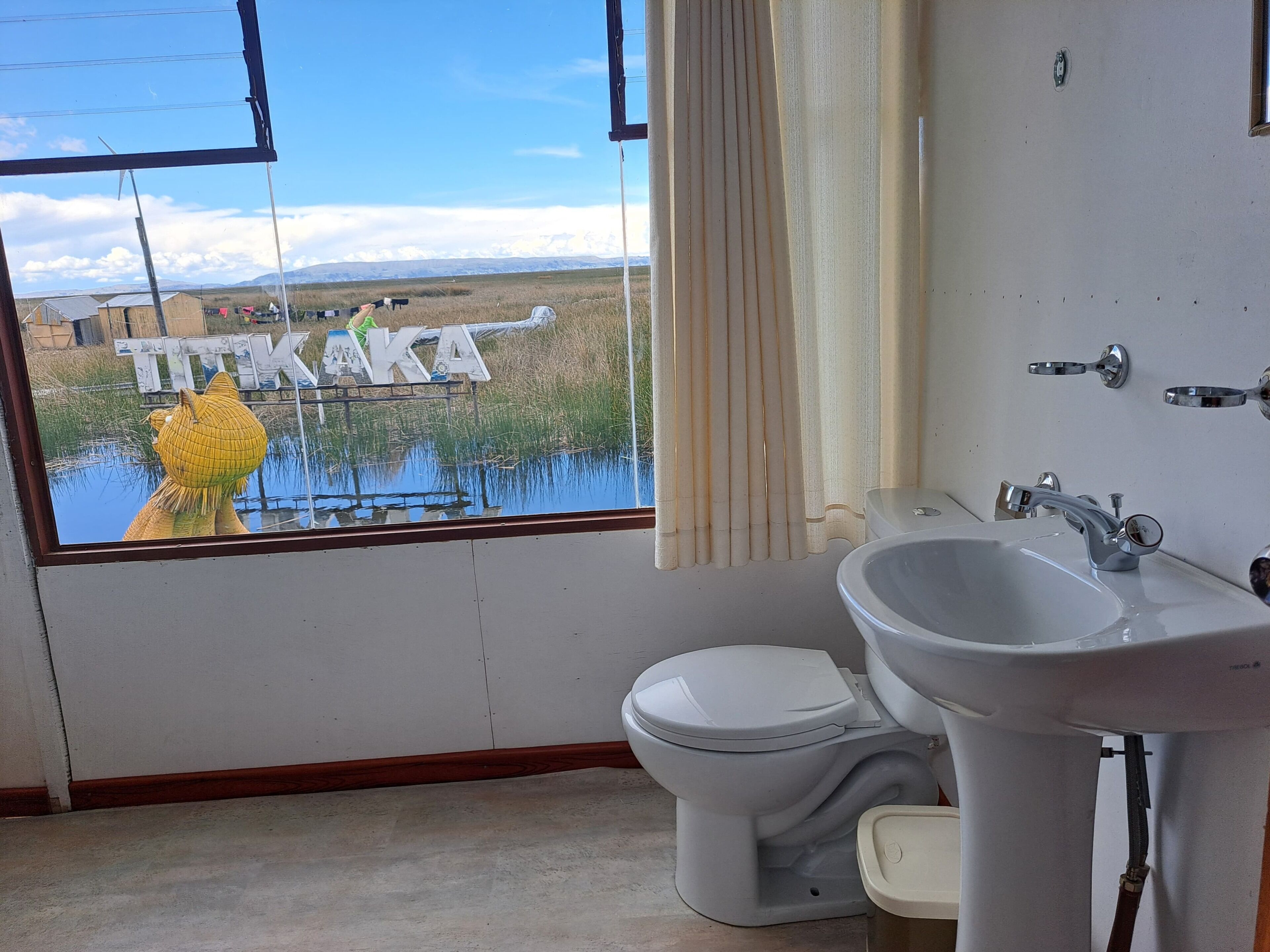 Casa de banho com sanita, lavatório e janela com vista para uma paisagem com um letreiro a dizer 'TEKAKA'.