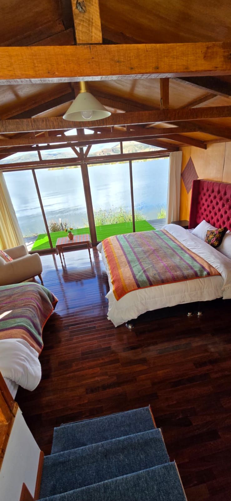 A wooden cabin bedroom with a bed, a chair, a table, and a view of the lake.
