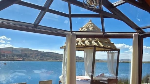 A room with a large glass window, wooden flooring, a sofa, a chair, a small table, and a decorative item. - Uros Kaivalia Lodge (Puno)
