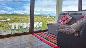 A room with a large window, a sofa with colorful pillows, and a view of a field with a sign. - Uros Kaivalia Lodge (Puno)