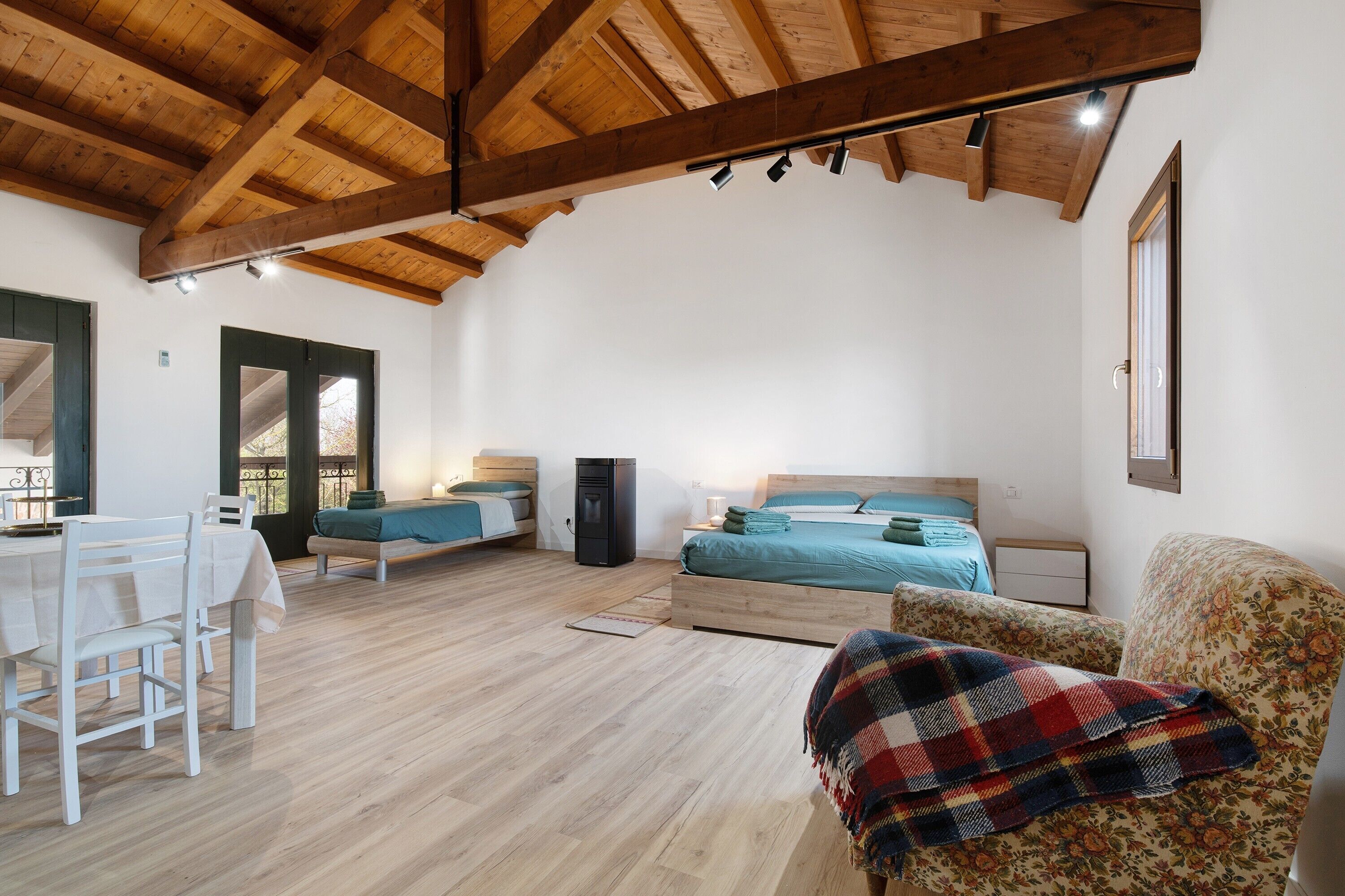 A spacious room with wooden beams, a bed with blue bedding, a desk with chairs, and a floral armchair.