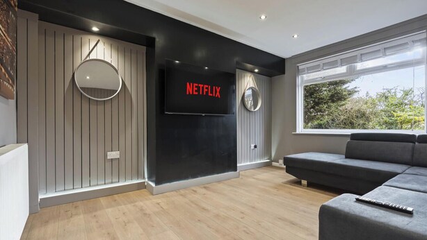 Uma sala de estar moderna com uma grande televisão de tela plana presa a uma parede preta, um espelho redondo e um sofá seccional cinza.