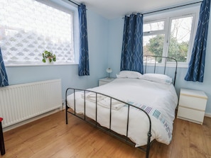 Un dormitorio con estructura de cama metálica, ropa de cama blanca, una mesita de noche con lámpara, un radiador y una ventana con cortinas azules.