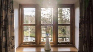 A window with a view of trees and a body of water, a white vase with branches, and a wooden window frame. - Wharf Cottage: Swimming lake, BBQ Hut, Games Room (Alton Towers & Staffordshire Villages)