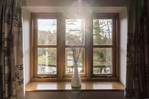 Une fenêtre donnant sur des arbres et un plan d’eau, un vase blanc orné de branches et un cadre de fenêtre en bois.
