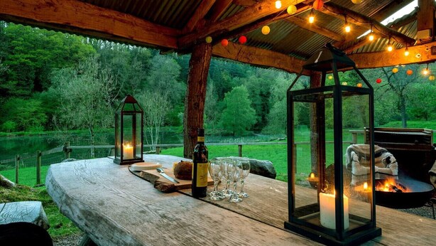 An outdoor dining area with a rustic wooden table, lanterns, and a fire pit. - Wharf Cottage: Swimming lake, BBQ Hut, Games Room (Alton Towers & Staffordshire Villages)