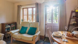 A cozy living room with a sofa, a dining table with plates and a wine glass, a bookshelf, and a window with curtains. - Wharf Cottage: Swimming lake, BBQ Hut, Games Room (Alton Towers & Staffordshire Villages)