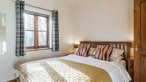 A bedroom with a bed, curtains, a window, a bedside table, and a door. - Clinkum Cottage Swimming Lake, BBQ hut, Games Room (Alton Towers & Staffordshire Villages)