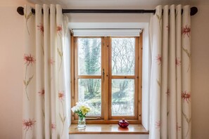 Une fenêtre ornée de rideaux à motifs floraux, un cadre de fenêtre en bois et un vase rempli de fleurs posé sur le rebord de la fenêtre.