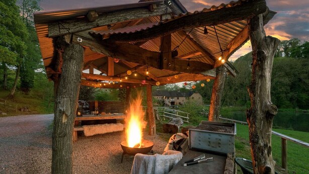 A rustic outdoor seating area with a fire pit, wooden tables, and string lights. - Bolt Hole: Swimming Lake, BBQ hut, Games Room (Alton Towers & Staffordshire Villages)