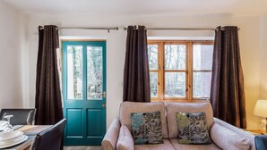 A cozy room with a teal door, a sofa with patterned cushions, a dining table with plates, and a window with curtains. - Wysteria: Swimming Lake, BBQ hut, Games Room (Alton Towers & Staffordshire Villages)