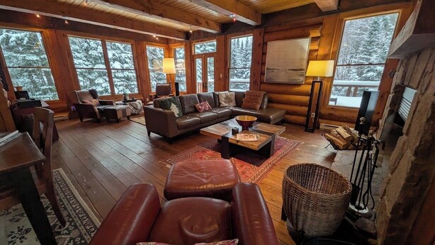 L’intérieur d’une cabane en rondins, avec un plancher en bois, des canapés en cuir, une table basse et de grandes fenêtres donnant sur un paysage enneigé.