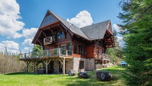 Une cabane en rondins dotée d’un balcon, avec des fondations en pierre et une vue sur la forêt environnante.