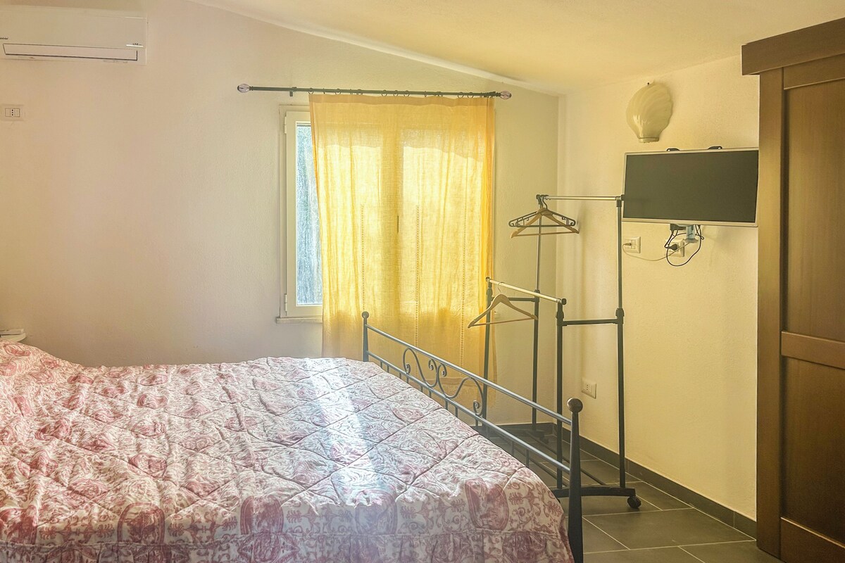 Een slaapkamer met een bed, een raam met gordijnen, een kledingrek en een televisie aan de muur.