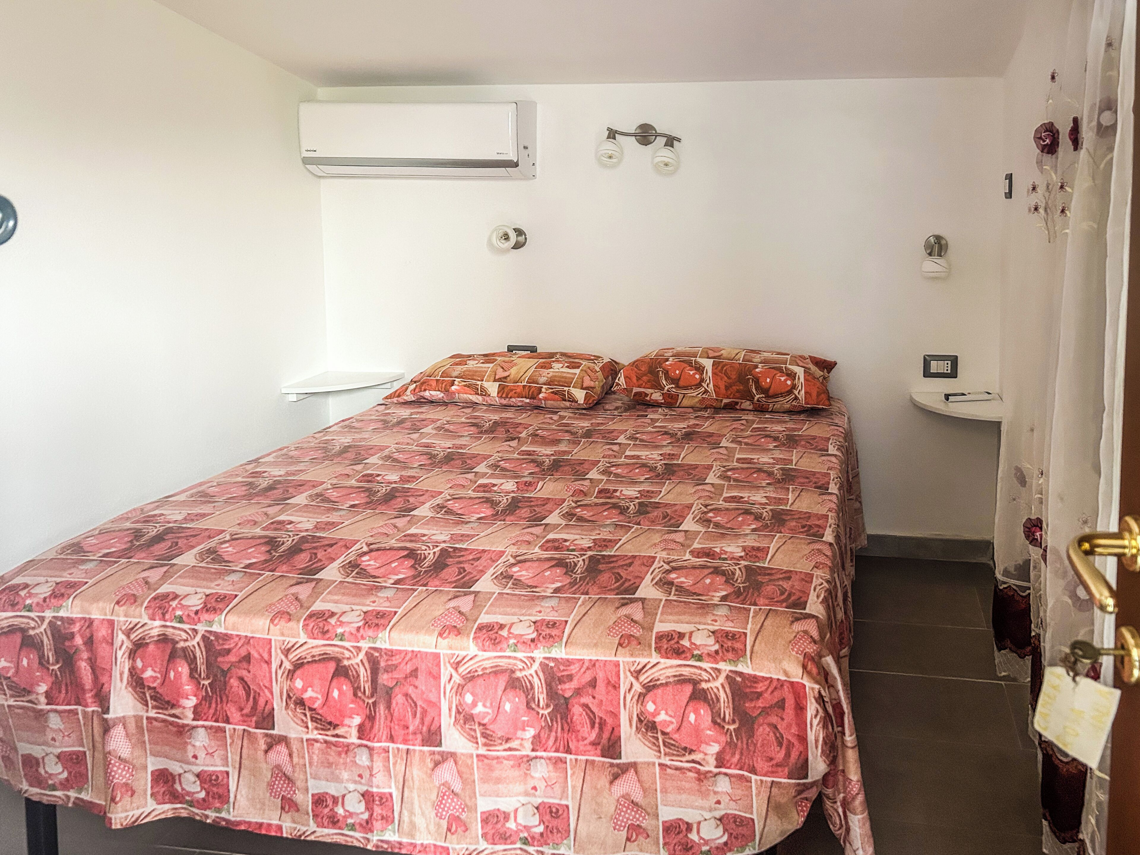 Une chambre à coucher comprenant un lit, un climatiseur, un luminaire fixé au mur, une petite table de chevet et un rideau.