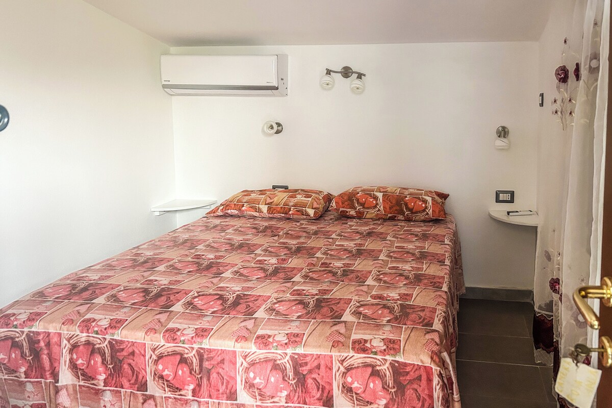 Een slaapkamer met een bed, een airconditioner, een wandlamp, een klein nachtkastje en een gordijn.