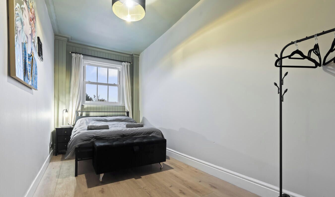A bedroom with a bed, a bedside table, a window with curtains, a floor lamp, and a coat rack.