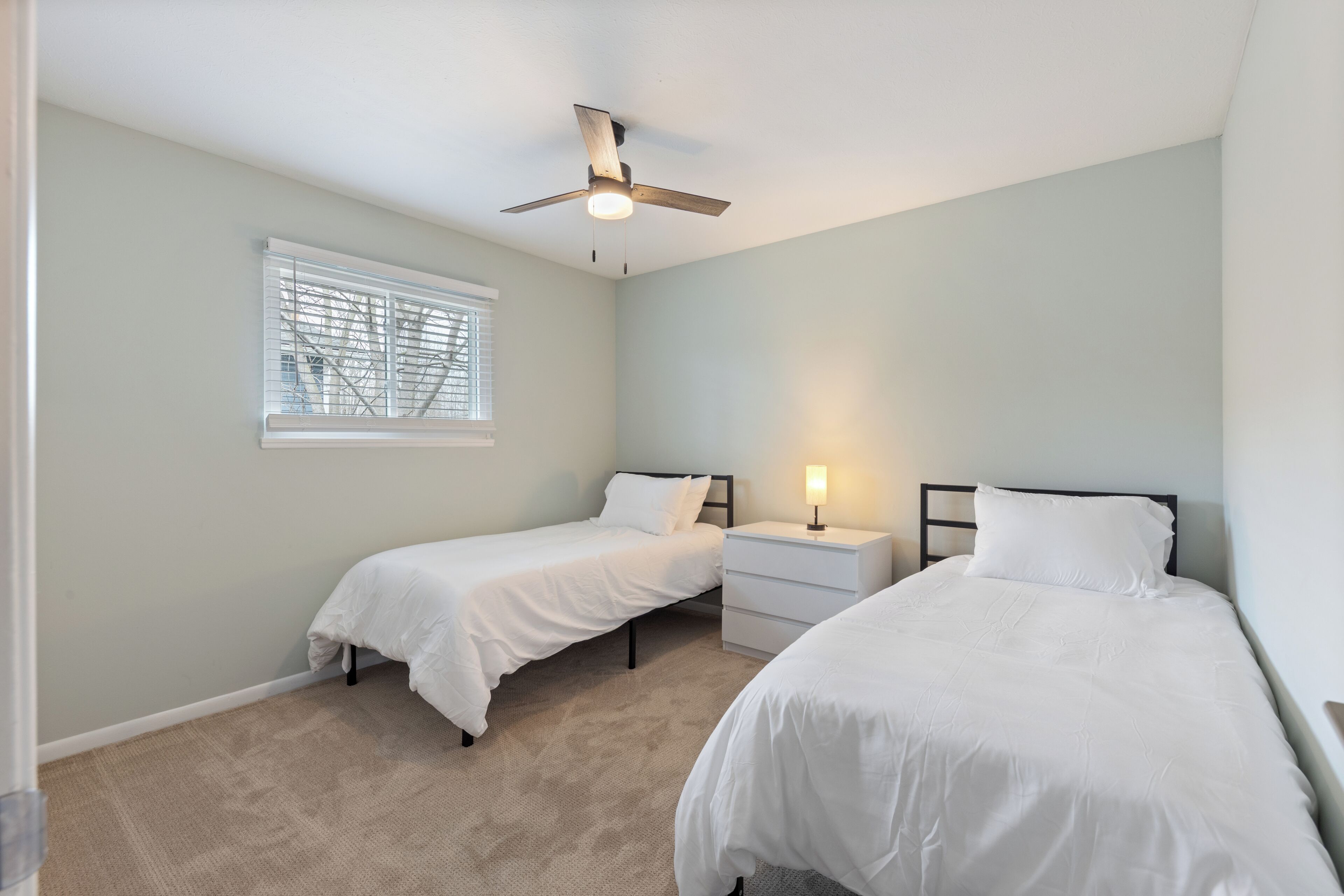 A bedroom with two single beds, a ceiling fan, a nightstand with a lamp, and a window with blinds.