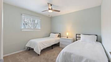 A bedroom with two single beds, a ceiling fan, a nightstand with a lamp, and a window with blinds.