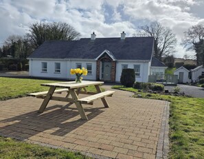 A house with a paved patio, a wooden picnic table with a vase of flowers, and a well-maintained lawn.