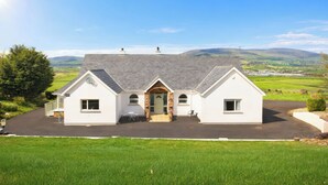 A two-story house with a brick entrance, a driveway, and a view of the countryside. - An Grianán Getaway (Burt)