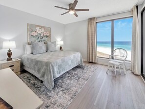 A bedroom with a bed, a nightstand, a chair, a ceiling fan, and a large window with a view of the beach. - Beach Break  3 bed 3 bath (Navarre Beach)